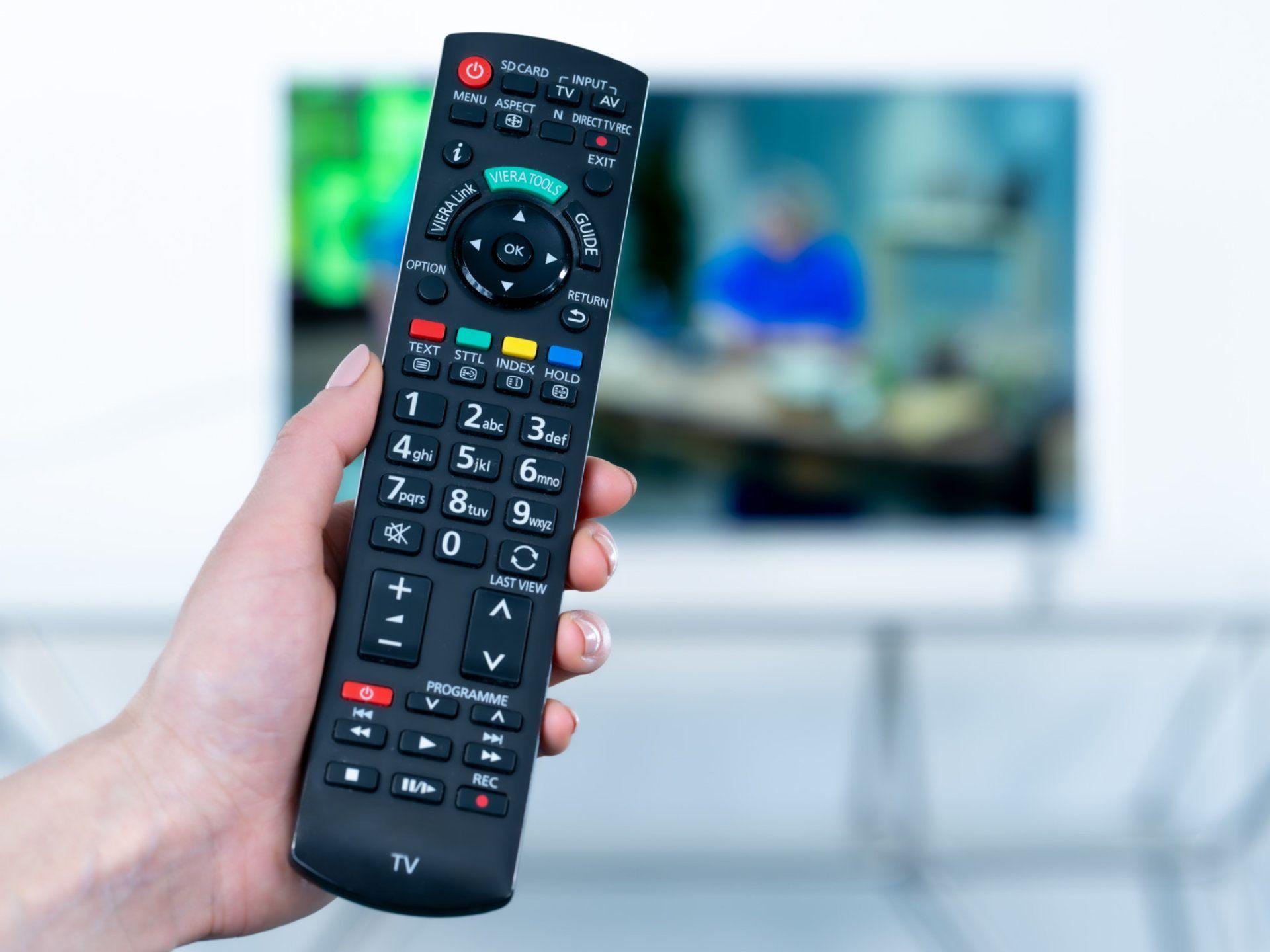 Hand hält eine Fernbedienung, im Hintergrund ist ein eingeschalteter Fernseher zu sehen.